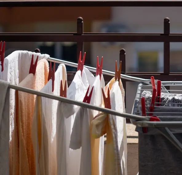 Zdjęcie przedstawia ubrania rozwieszone na suszarce balkonowej. Część prania składa się z jasnych materiałów, takich jak ręczniki i białe koszule, które zostały przymocowane do sznurków za pomocą czerwonych klamerek. Ubrania delikatnie powiewają, co sugeruje obecność lekkiego wiatru. W tle widoczna jest balustrada balkonu oraz częściowo rozmazana fasada budynku, wskazująca na miejskie otoczenie.Po prawej stronie widoczna jest także część innej suszarki, na której znajdują się szare i wzorzyste elementy odzieży. Całość ma przyjemny, codzienny charakter, podkreślając troskę o świeżość ubrań i dbałość o garderobę.